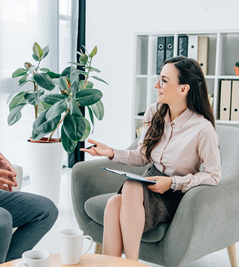 woman with glasses sits in a chair and with a clipboard and talks to the person in front of her