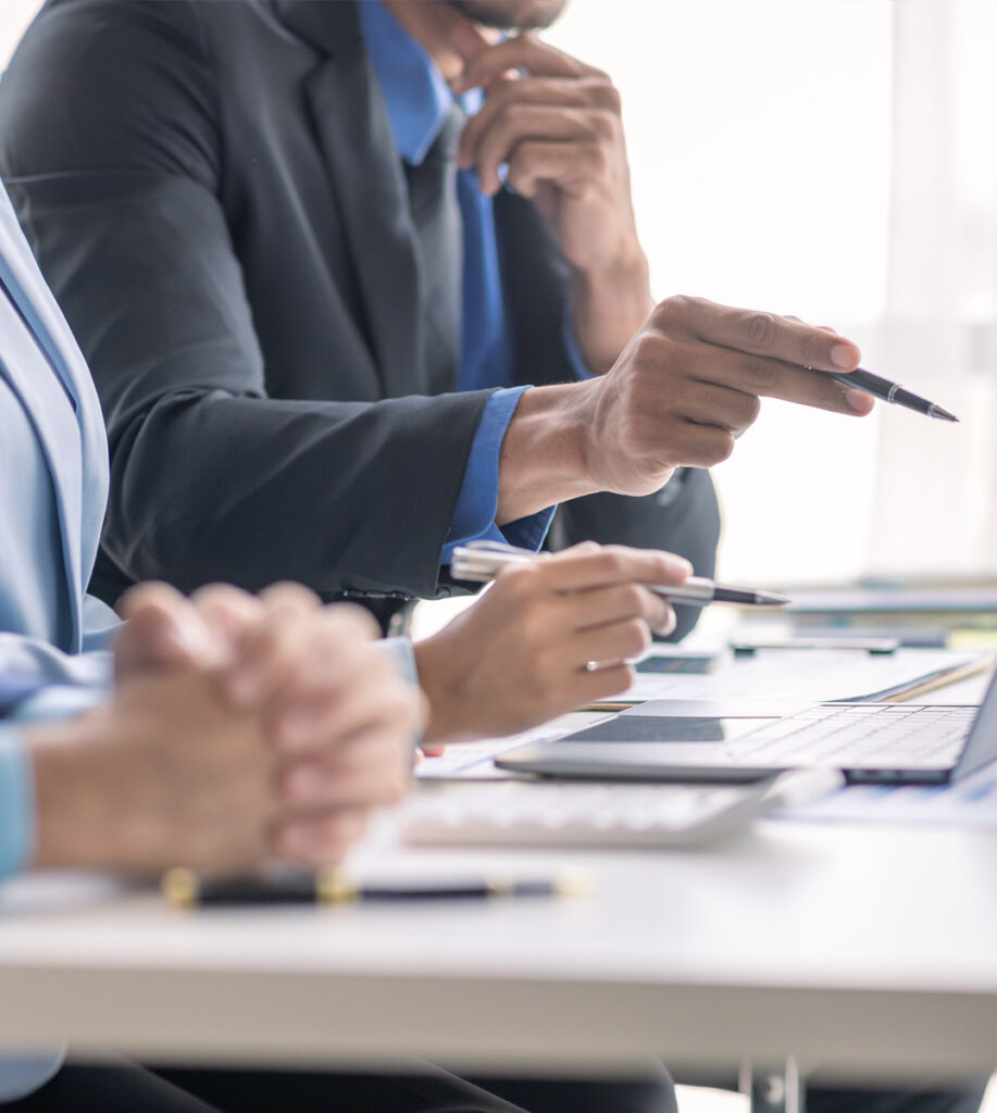 people sit at meeting table and one person points at the screen of a laptop