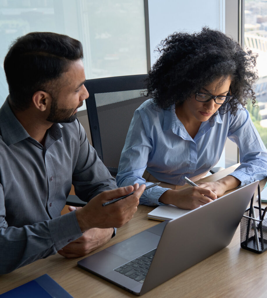 woman with glasses takes notes as a man is talking to her as they sit at a meeting table and laptop in front of them