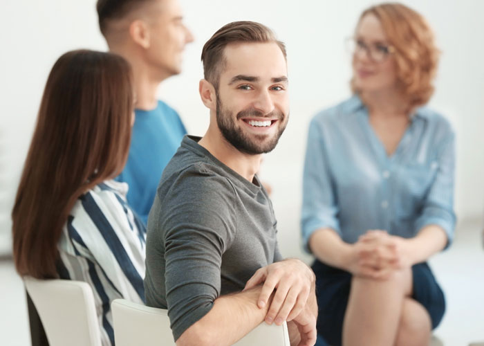 man sits and smiles as other people in the background talk together